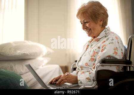 Sorridente donna senior divertirsi usando un computer portatile mentre è seduto in una sedia a rotelle. Foto Stock