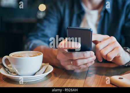 L'uomo le mani in denim shirt scorrere con il dito sullo schermo il suo smart phone, vicino a tazza con il caffè sul tavolo in legno Foto Stock