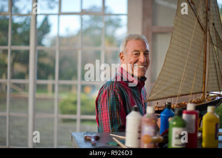 Ritratto di un sorridente senior uomo con un modello di barca dentro il suo garage. Foto Stock