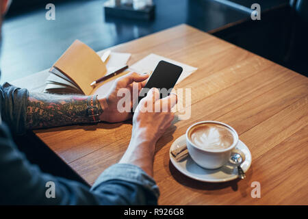 L'uomo le mani in denim shirt scorrere con il dito sullo schermo il suo smart phone, vicino a tazza con il caffè sul tavolo in legno Foto Stock