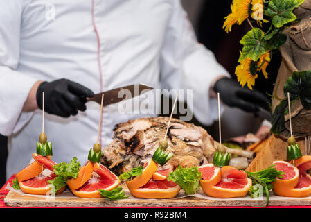 Spettacolo al ristorante: il capo cuoco tagli in pezzi appena cucinata coscia di maiale, il processo da vicino. Decorazioni di frutta e verdura sulla tavola. Foto Stock