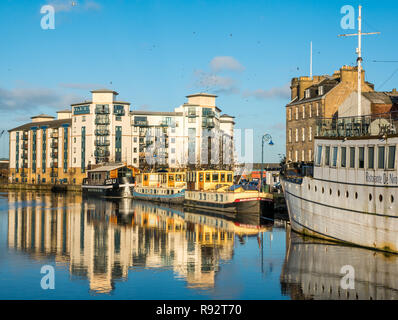 Leith, Edimburgo, Scozia, Regno Unito. Xix Dec, 2018. Regno Unito Meteo: una bella e soleggiata giornata invernale nella capitale con la mancanza di vento creando riflessi nell'acqua del fiume di Leith. Queen's Quay, un moderno appartamento sviluppo si riflette nell'acqua come pure numerosi fiume chiatte o case galleggianti Foto Stock