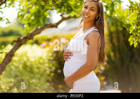 Sorridente donna incinta in piedi in giardino tenendo le mani sulla pancia Foto Stock