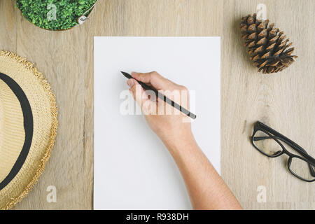 La mano dell'uomo detiene una scritta a matita su carta A4 posto su una scrivania in legno con vasi di piante, Pigna, cappello e occhiali. Foto Stock