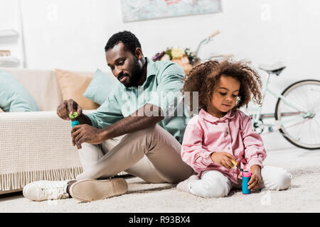 African American padre e figlia seduti sul pavimento, rendendo le bolle di sapone e guardando verso il basso Foto Stock