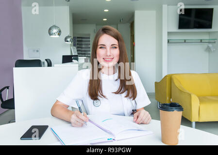 Ritratto di giovane donna sorridente medico rendendo note in notebook mentre seduto dietro il tavolo con telefono e caffè all'interno della clinica medica. Studen Foto Stock