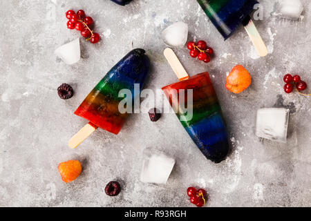 Rainbow coloratissimo gelato con lampone giallo e rosso, blackberry, ribes ghiaccioli con bastone di legno su sfondo grigio. Vista dall'alto. Sfondo di cibo Foto Stock