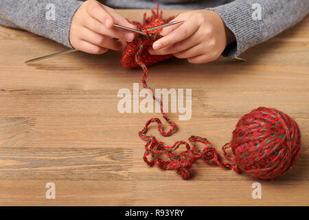 La ragazza del bambino è quello di imparare a lavorare a maglia una sciarpa. Rosso di filati di lana è sul tavolo di legno. Mano closeup. Foto Stock