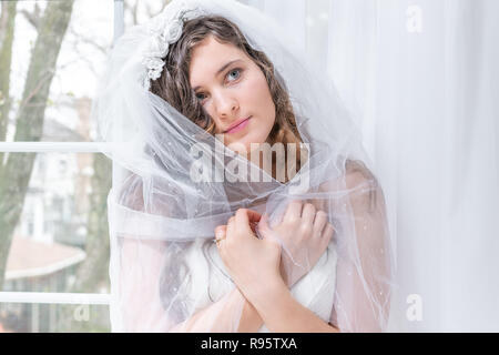 Closeup Ritratto di giovane donna uomo donna, sposa in abito da sposa, azienda, avvolgente, coprendo circa le spalle con velo avvolto, dalla finestra, whi Foto Stock