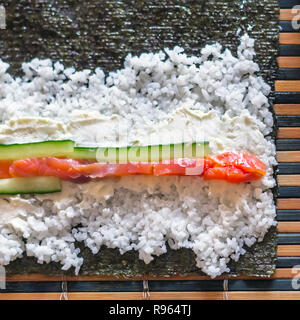 Effettua il Sushi rotoli: Salmone Pesce strisce, bastoncini di cetriolo, crema di formaggio, riso e Nori alghe foglio su un tappetino di bambù Foto Stock