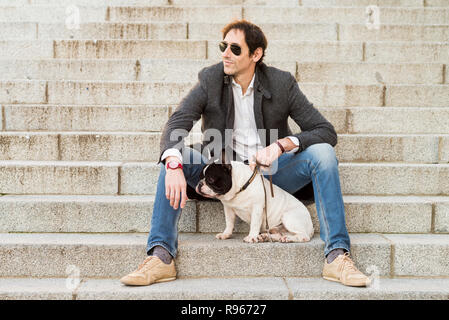 Scena urbana. Uomo vestito in abiti casual seduto su alcune scale posa vicino al suo cane Foto Stock