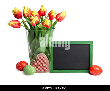 Lavagna con lo spazio di testo, mazzetto di rosso striata tulipani in vaso di vetro, uova di Pasqua e il cuore di legno isolato su sfondo bianco Foto Stock