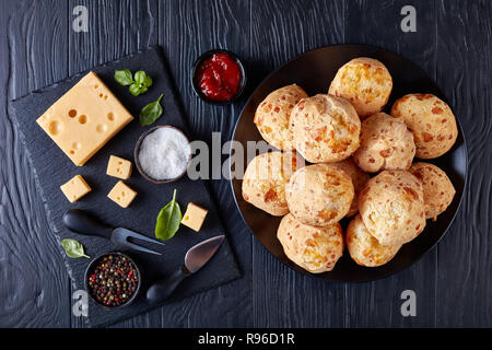 Gougeres, formaggio bignè palle su una piastra nera. Tradizionale formaggio francese choux ciambelle. Ingredienti e salsa di pomodoro su tavoli in legno nero, vista da un Foto Stock