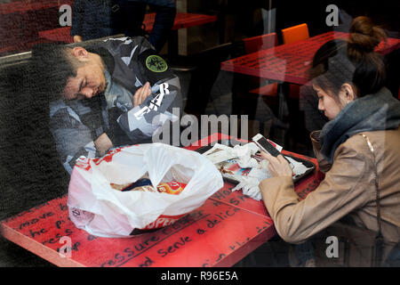 Uomo asiatico addormentato in KFC mentre il suo partner utilizza il suo smartphone, Barcelona, 2018. Foto Stock