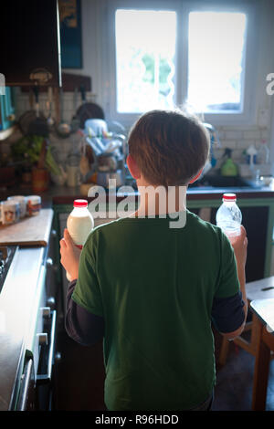 Vista posteriore del ragazzo di 11 anni in piedi in una cucina, circa per preparare tazze di tè, in grado di contenere fino a due bottiglie di plastica del latte nella sua mano sinistra e mano destra Foto Stock