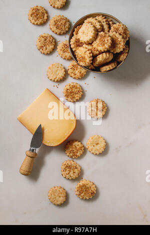 Snack caserecci frollini formaggio biscotti di sesamo nella ciotola e pezzo di formaggio a pasta dura con un coltello su marmo bianco dello sfondo. Laici piana, spazio Foto Stock