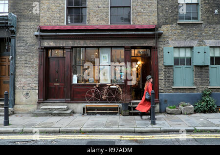 Donna in cappotto arancione oltrepassando negozio di antiquariato con bicicletta su Fournier Street vicino a Spitalfields Market in East End di Londra UK KATHY DEWITT Foto Stock