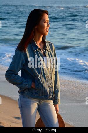 Una giovane donna, che indossa una giacca di denim e jeans, sta godendo di una pacifica nel tardo pomeriggio passeggiata sulla spiaggia. Foto Stock