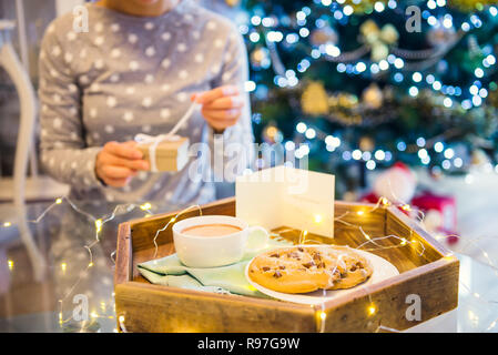 Natale e Anno Nuovo composizione. Cacao e biscotti sul vassoio in legno e sfocata senza volto della donna la sua apertura presente su sfondo di Natale. Dolci per Foto Stock