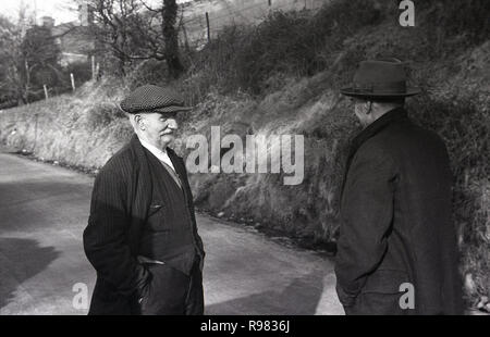 1940s, storico, due minatore gallese uno indossando un tappo piatto, altri un cappello, in piedi a una conversazione sulla strada di un paese al di fuori di Merthyr, Galles. Foto Stock