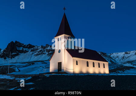 Ben illuminata di Vik chiesa si erge drammaticamente sulla città di Vik, in inverno in Islanda Foto Stock