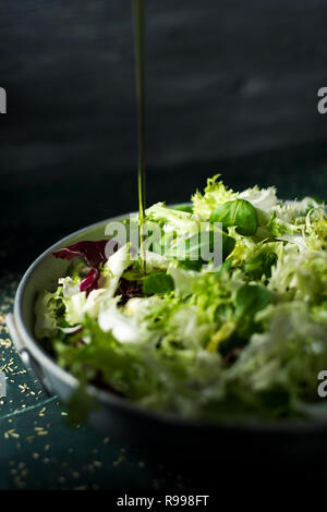 Primo piano di un fiotto di olio di oliva a cadere su di un mix di diverse foglie di insalata, come lattuga romana, indivia o rucola, su un rustico di una capsula metallica Foto Stock