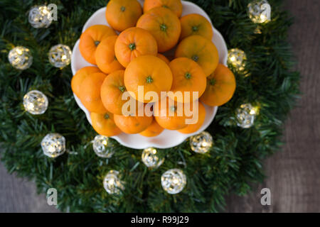 Pila di arance satsuma su una piastra bianca, in una corona di fiori con LED luci di Natale su uno sfondo di legno Foto Stock
