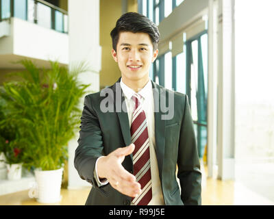 Giovani asiatici corporate executive raggiungere fuori per una stretta di mano, guardando la telecamera sorridendo. Foto Stock