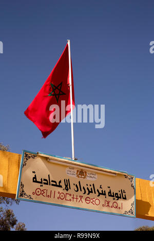 Il Marocco, Errachidia Provincia, Rissani, bandiera nazionale al di sopra della scuola segno di gate in arabo e script Berbera Foto Stock