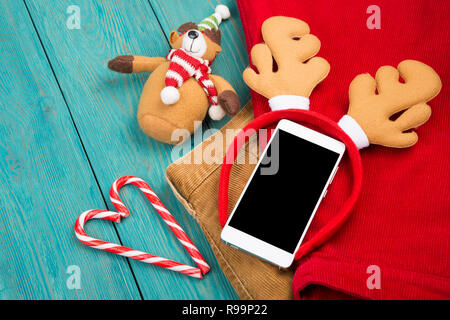 Giocattolo corna di renne, smartphone, Teddy bear, candy e vestiti blu sulla scrivania in legno Foto Stock