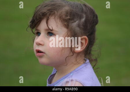 Giovane bruna bambino con grandi occhi blu che guardano lontano dalla macchina fotografica su uno sfondo di erba Foto Stock