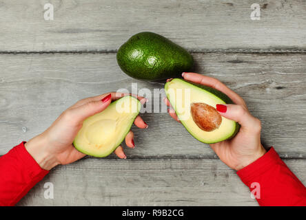 Vista del piano portapaziente, donna mano che regge due metà di avocado in ogni mano, ancora una pera intera collocato su grigio scrivania in legno sopra. Foto Stock