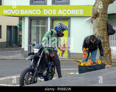 Moto Rider con il casco in testa si arresta per l'acquisto di un mazzo di fiori da un maschio di venditore ambulante davanti a 'Nuova Banca delle Azzorre " Foto Stock