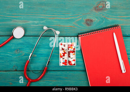 Luogo di lavoro di un medico. Red stetoscopio, notepad e pillole blu sulla scrivania in legno Foto Stock