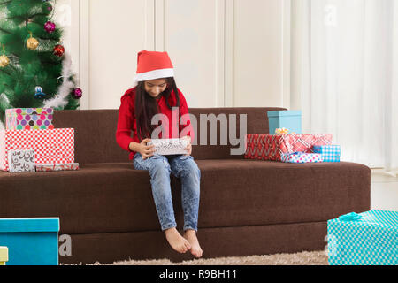 Piccolo felice carina ragazza con un regalo di Natale scatola seduti sul divano nella decorazione soggiorno con cappello a Natale Foto Stock