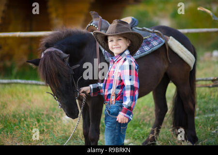 Carino fermer bello ragazzo cowboy in jeans godendo di giorno di estate in vita del villaggio con fiori in pelle da indossare mucca hat happyly sorridente e seduta sul cavallo Foto Stock