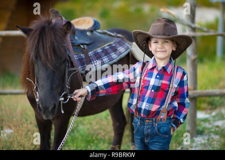 Carino fermer bello ragazzo cowboy in jeans godendo di giorno di estate in vita del villaggio con fiori in pelle da indossare mucca hat happyly sorridente e seduta sul cavallo Foto Stock