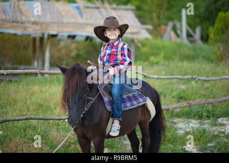 Carino fermer bello ragazzo cowboy in jeans godendo di giorno di estate in vita del villaggio con fiori in pelle da indossare mucca hat happyly sorridente e seduta sul cavallo Foto Stock