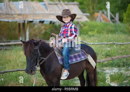 Carino fermer bello ragazzo cowboy in jeans godendo di giorno di estate in vita del villaggio con fiori in pelle da indossare mucca hat happyly sorridente e seduta sul cavallo Foto Stock