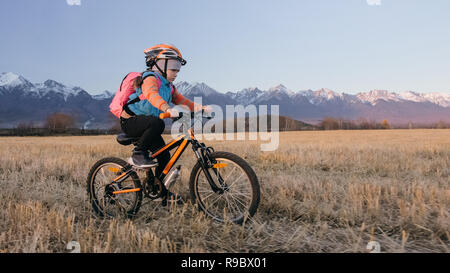 Uno caucasico giostre per bambini bici nel campo di grano. Bambina a cavallo nero ciclo arancione su sfondo di belle montagne innevate. Biker moto giro con zaino, casco. Mountain bike hardtail. Foto Stock