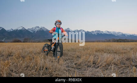 Uno dei bambini caucasici a piedi con bici nel campo di grano. Poco ragazza camminare nero ciclo arancione su sfondo di belle montagne innevate. Biker stand con zaino e casco. Foto Stock