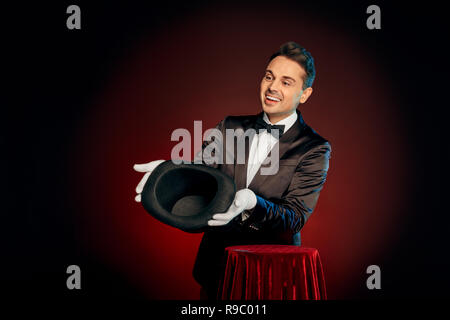 Professional mago indossare tuta e guanti isolati permanente su nero e sfondo rosso rendendo trucco mostra vuoto top hat al pubblico sorridente ven Foto Stock