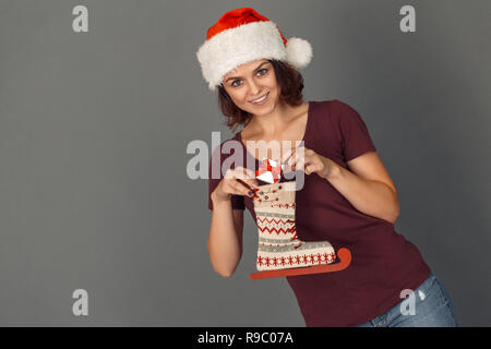 Giovane donna indossa santa hat tema natalizio isolato in piedi sul muro grigio messa piccola scatola regalo decorata in skate boot cerca fotocamera a sorridere h Foto Stock