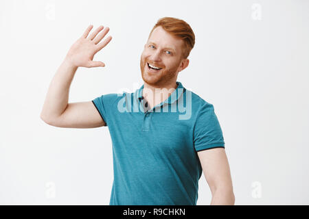 Bello felice redhead uomo in attesa di alta cinque, sorridente largamente il sollevamento palm alta per salutare la persona in modo moderno e accogliente vicino uno su uno sfondo grigio, indossando trendy Polo shirt Foto Stock