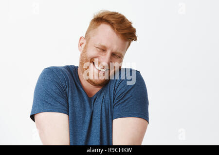 Oh arrestarlo. Ritratto di carino soddisfatti e contenti e felici divertente redhead uomo in blu t-shirt, scrollare e faccia pendente sulla spalla mentre sorridente largamente, arrossendo dalla gioia e felicità Foto Stock