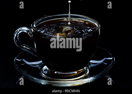 Gocce di latte che rientrano in un vetro trasparente con tazza di caffè nero su uno sfondo nero, studio luce Foto Stock