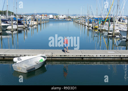 Marina con barche ormeggiate in calme acque blu e piccolo ragazzo in rosso giubbotto di salvataggio nel molo con canna da pesca. Foto Stock