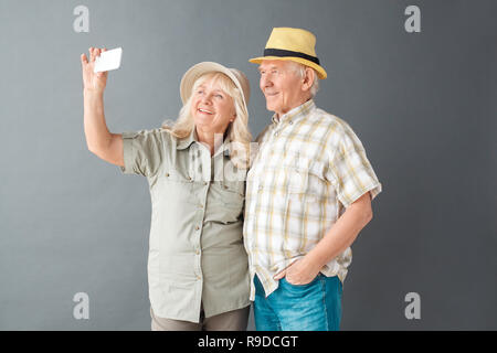 Coppia senior turisti spiaggia indossare cappelli studio isolato in piedi sul muro grigio tenuto selfie foto sullo smartphone guardando la fotocamera in posa sorridente joyfu Foto Stock