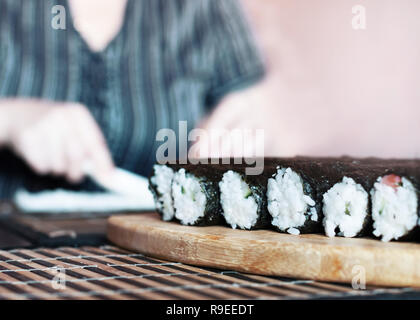 Effettua il Sushi rotoli a casa: rotoli tenuta sulla tavola di legno prima di affettatura. Donna sfocata mani diffondere il riso attraverso il foglio di Nori con plastica Sp Foto Stock