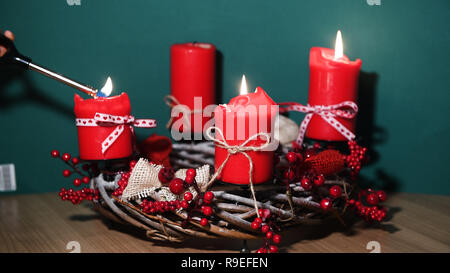 Le mani della madre e del bambino, tenendo in mano un accendino, accendendo candele sulle moderne ghirlanda di Natale con quattro candele rosse su una superficie di legno con verde Foto Stock